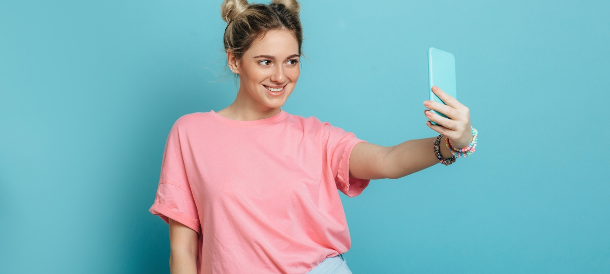 Social media : des bonnes pratiques aux mauvaises habitudes Une femme avec un t-shirt rose qui prend un selfie devant un fond bleu