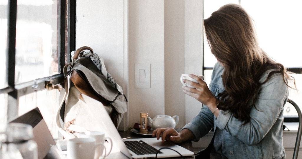 Une femme brune, porte une veste en jean. Elle se tient devant son bureau , sur son ordinateur et bois un café