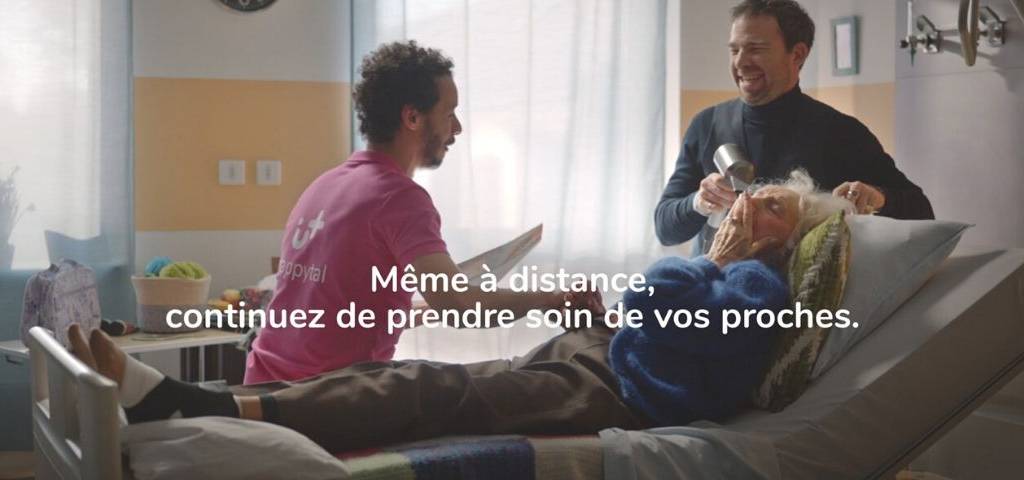 Homme assis sur un lit d'hopital en train de converser avec une femme agée pendant qu'elle se fait coiffer, le tout en riant