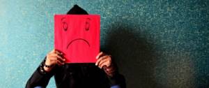 Personne assise par terre, en noir, tenant devant son visage une feuille A4 rouge avec les traits d'un visage triste