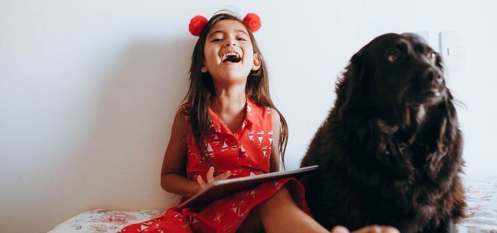 Petite riant avec une tablette dans les mains, assise sur son lit, accompagnée de son chien