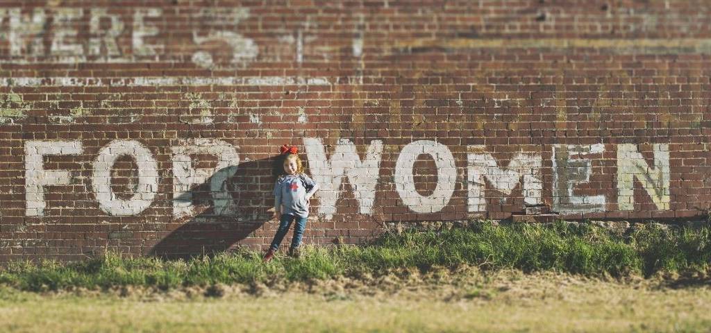 Petite fille devant un mur de briques rouges sur lequel est inscrit  - Women - 