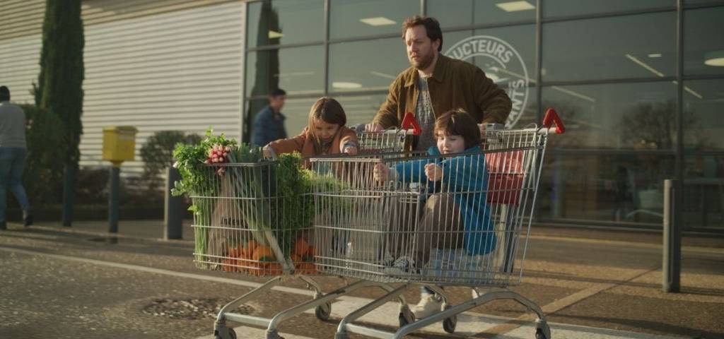 Papa sortant d'un supermaché Intermarché, avec 2 caddies, dans lesquels se trouvent un enfant qui fait mine de conduire une voiture