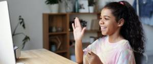 Jeune fille devant son ordianteur, levant la main pour dire bonjour à son enseignant en ligne