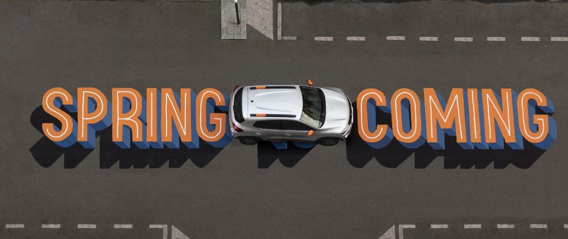Dacia célèbre le printemps dans la rue et clame haut et fort l’arrivée de Spring, la révolution électrique pour tous. Voiture Dacia prise en hauteur avec le message  - Spring is coming -  inscrit sur la route en orange et bleu