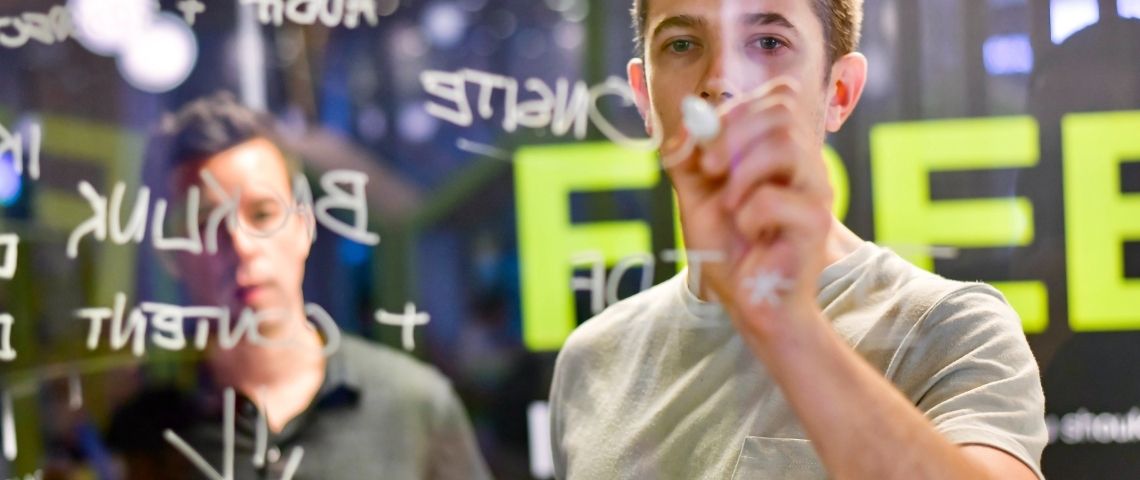 xcub-bouygues-telecom-lancement-deuxieme-edition-programme-flowers-numerique-start-up-innovation Un homme qui écrit sur un tableau transparent avec un autre homme derrière lui qui regarde