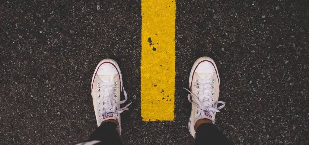 Des pieds. Chaussures : converses blanches. Bitume face une ligne de peinture jaune