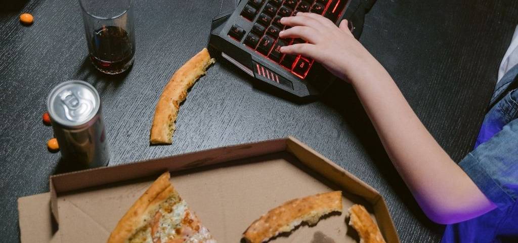 Main sur un clavier d'ordinateur, avec un verre de soda et une pizza entâmée posées à côté