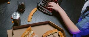 Main sur un clavier d'ordinateur, avec un verre de soda et une pizza entâmée posées à côté