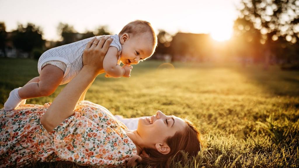 Maman portant son bébé au dessus delle allongée dans un pré
