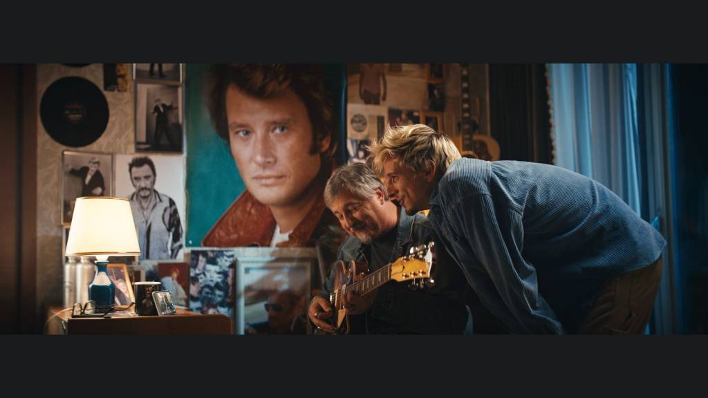 2 hommes, l'un jouant de la guitare, l'autre penché vers lui avec des posters de Johnny Hallyday en arrière plan