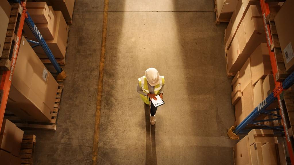 Vue descendante : Un travailleur portant un casque dur vérifie les stocks et les stocks à l’aide d’un ordinateur tablette numérique dans l’entrepôt de détail rempli d’étagères avec des marchandises