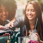 Femme faisant un paiement avec son smartphone à une terrasse entourée de ses amis