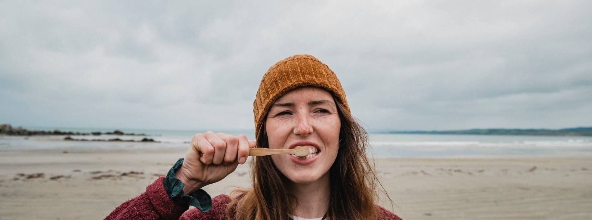 Quelles solutions pour lutter contre les 10kg de déchets que représentent les brosses à dents par personne et par an en France ? Femme se brossant les dents avec une plage en arrière plan