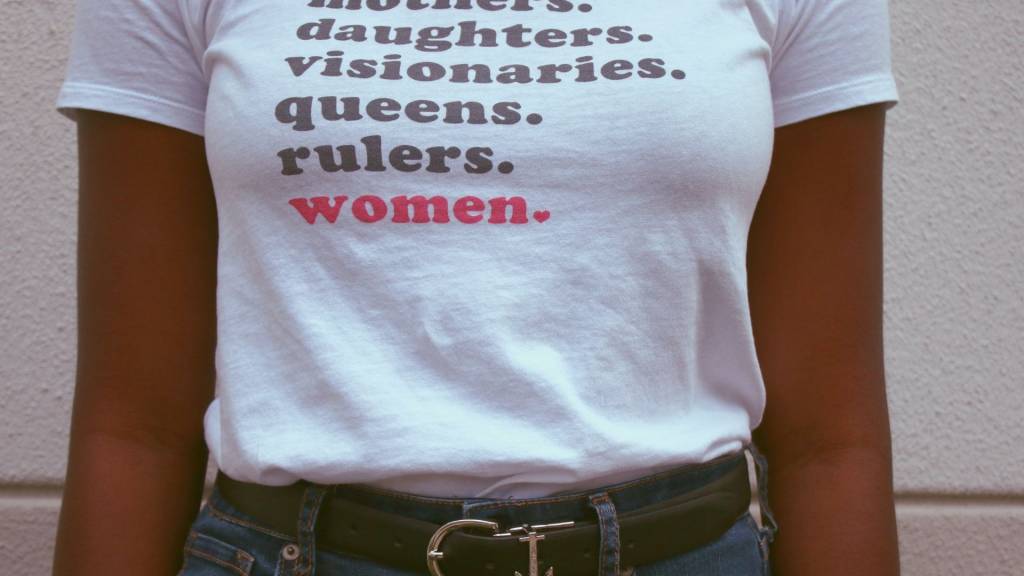 Femme avec un t-shirt avec les mots : women is rulers, queens, visionaries, daughters, mothers