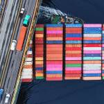 Porte contenaire passant sous un pont sur lequel se trouvent des camions